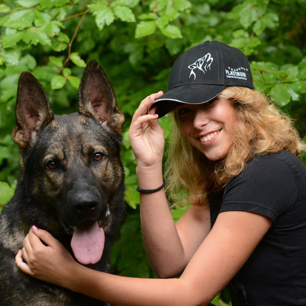 Ženska s kodrastimi svetlimi lasmi, ki nosi Ekskluzivna PLATINUM kapa s šiltom, se nasmiha ob srečnem nemškem ovčarju sredi zelenega listja - idealen navdih za ljubiteljske dodatke za kosmatince znamke PLATINUM.