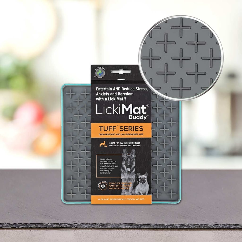 Podloga LickiMat® - TUFF BUDDY blagovne znamke PLATINUM stoji na mizi. Siva podloga, ki je zasnovana za zmanjševanje tesnobe psov, ima na površini dvignjen vzorec križa. Povečan krog prikazuje teksturo; na embalaži sta prikazana dva psa.