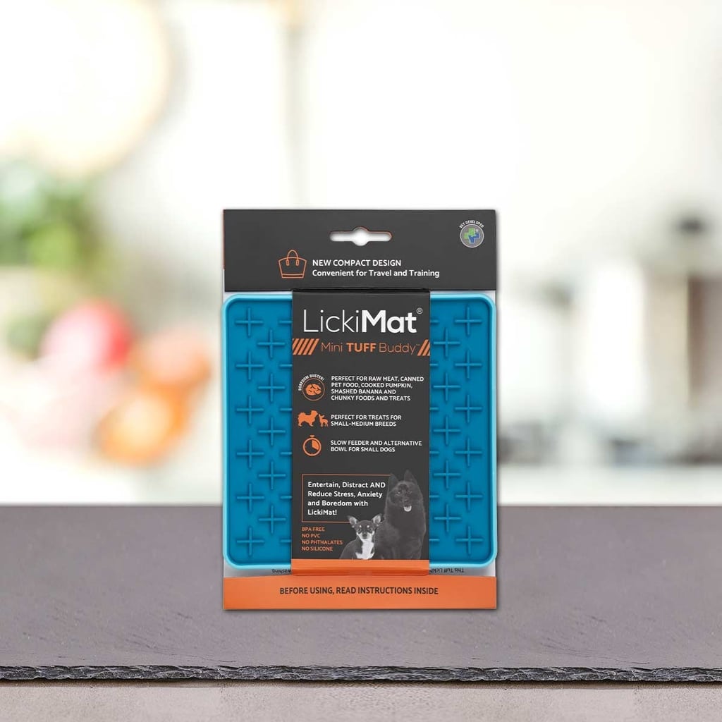 Modra PLATINUM Podloga LickiMat® - TUFF BUDDY v embalaži stoji pokonci na sivi površini z zamegljenim notranjim ozadjem. Na embalaži so navedene prednosti, kot je zmanjšanje tesnobe psa za pse in mačke.