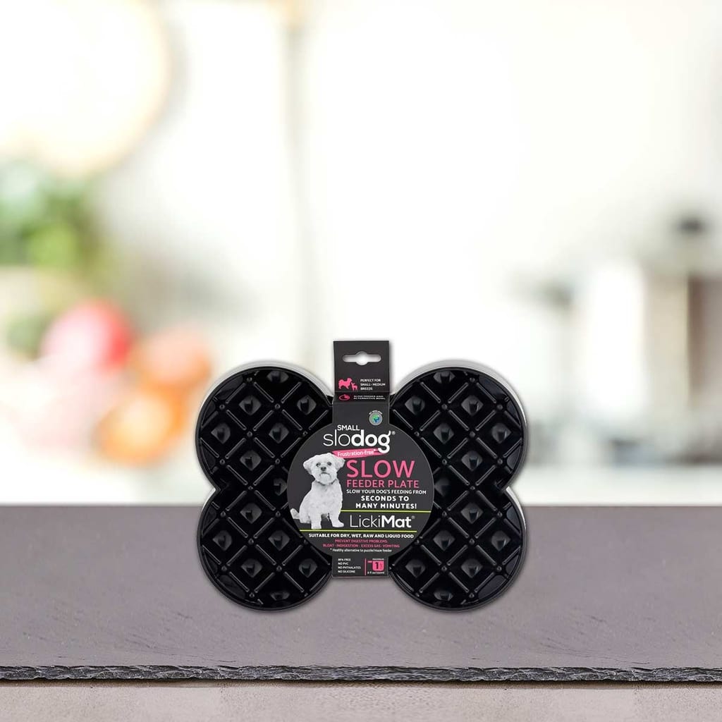 Črna plošča za počasno hranjenje v obliki kosti z napisom "Podloga LickiMat® - SLODOG" blagovne znamke PLATINUM, prikazana pokonci na kuhinjskem pultu z zamegljenim ozadjem; idealna za počasno hranjenje psov kot LickiMat ali pasja podloga za lizanje.
