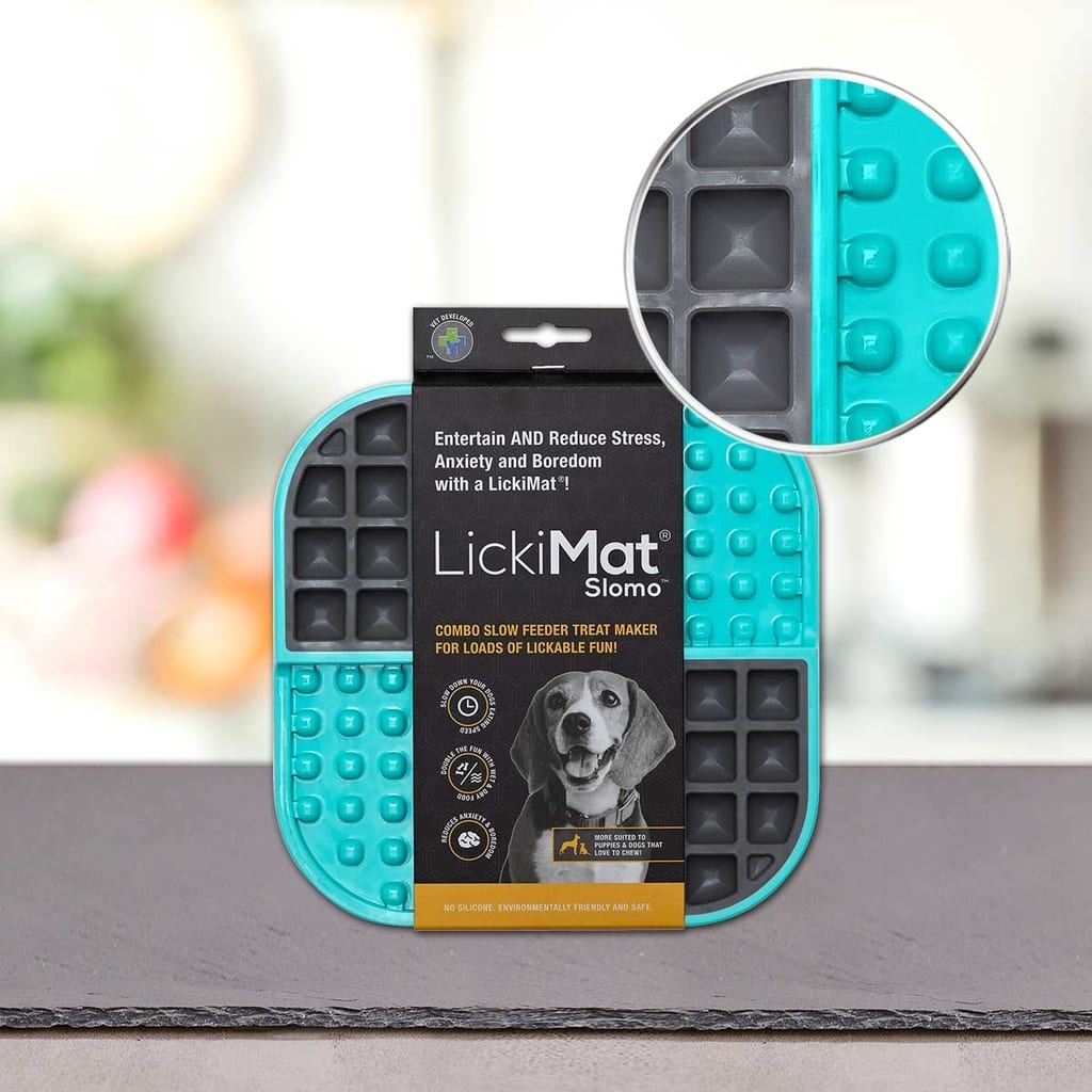 Na mizi je prikazana modra in siva PLATINUM Podloga LickiMat® - SLOMO. Na embalaži je nasmejan pes, ki promovira teksturirano površino podloge za upočasnjeno hranjenje, ki pomaga zabavati in umiriti hišne ljubljenčke.