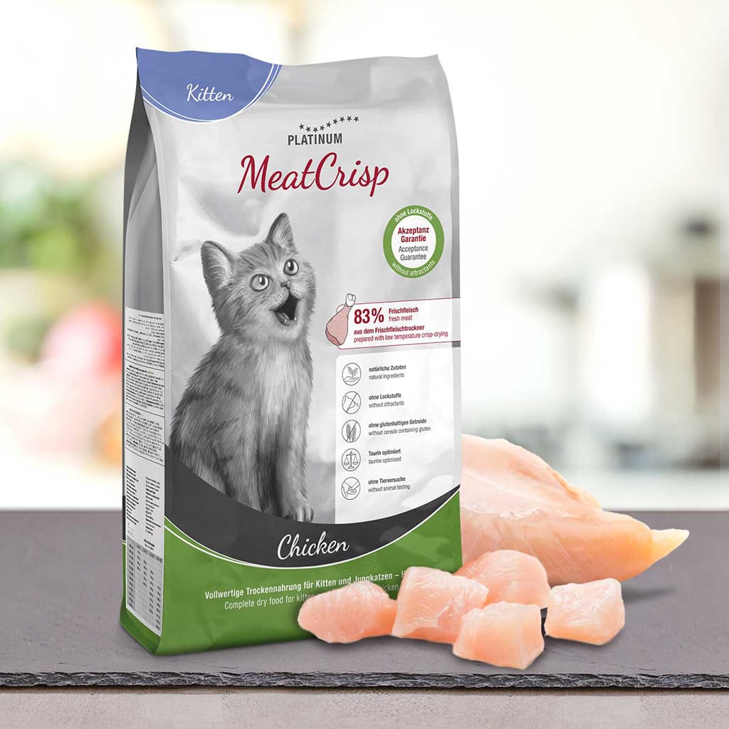 Vrečka PLATINUM MeatCrisp Kitten Chicken, Suha Hrana za Mačke stoji ob surovem piščancu na pultu. Na embalaži je prikazan siv maček, podatki o hranilni vrednosti in omenjen poseben postopek Freshmeatdryer.