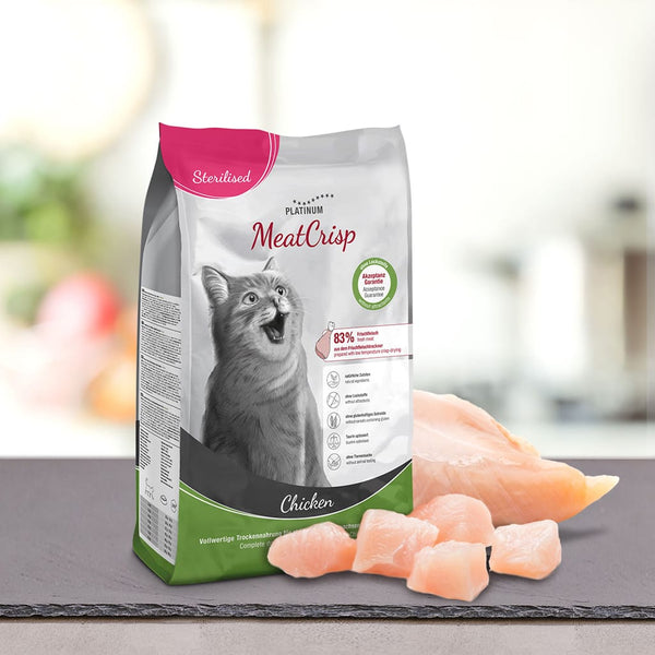 Vrečka PLATINUM MeatCrisp Sterilised Chicken, Suha Hrana za Mačke, s sivo etiketo Cat in Chicken, stoji za kosi sveže meso na temni površini.