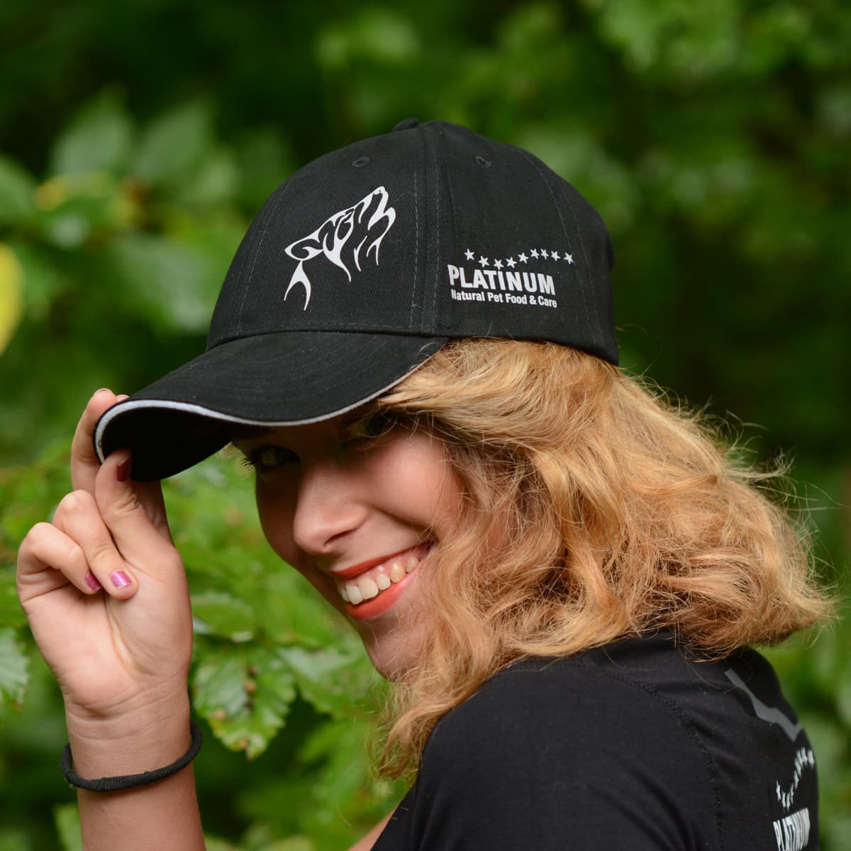 Nasmejana oseba z valovitimi svetlimi lasmi nosi kapo Ekskluzivna PLATINUM kapa s šiltom-tvoja nagrada za zvestobo!-izdelek PLATINUM z grafiko volka in besedilom, ki stoji na prostem med zelenim listjem.