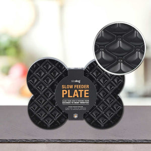 Na mizi je prikazana črna PLATINUM Podloga LickiMat® - SLODOG plošča za počasno hranjenje psov s teksturirano površino, ki je prikazana v bližnjem posnetku. Na nalepki je napis "Plošča za počasno hranjenje", spodaj pa so prikazana navodila; idealna kot pasja podloga za lizanje.