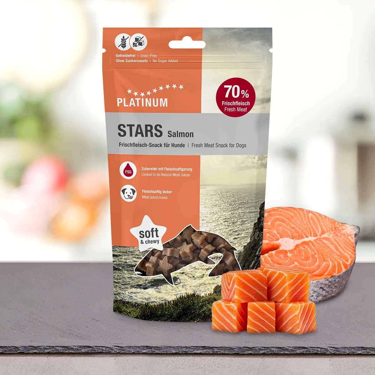 Vrečka PLATINUM Salmon Stars 185 g - nagrada iz svežega lososa, ki jo bo tvoj pes oboževal - stoji na mizi poleg koščkov surovega lososa in razgleda na obalo, kot nalašč za nagrajevanje psa s kakovostnimi priboljški.