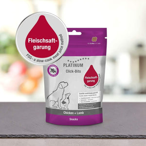 Na pultu je razstavljeno 150-gramsko pakiranje PLATINUM Click-Bits Chicken + Lamb Fit priboljški za Pse z vijoličasto in belo embalažo s psom, kužkom in rdečo etiketo Fleischsaftgarung.