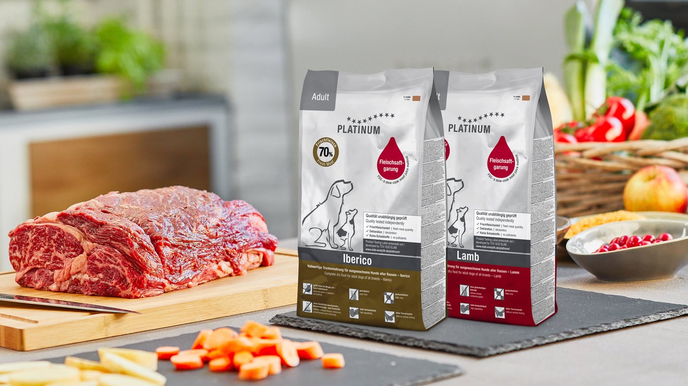 Na kuhinjskem pultu je lesena deska za rezanje s surovo govejo pečenko in narezanim korenjem. Zraven sta dve vrečki pasje hrane Platinum z oznakama Iberico in Lamb. V ozadju sta zelenjava in košara.