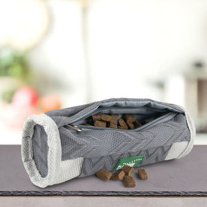 Na mizi je odprta siva torbica z zadrgo z napisom Paket okusov Priboljškov za Pse by PLATINUM, v kateri so rjavi pasji priboljški, nekaj pa se jih razlije. Rahlo zamegljeno ozadje daje izdelku večjo ostrino.